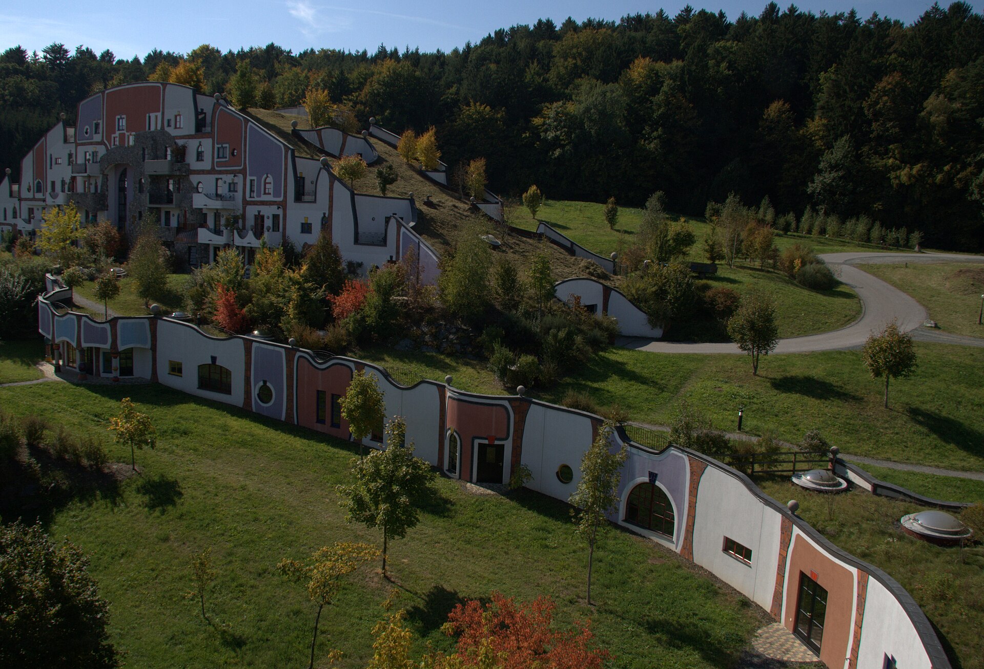 Aerial view of Rogner Bad Blumau: grass-roofed buildings with wavy rooflines set into a green hillside, forest behind.